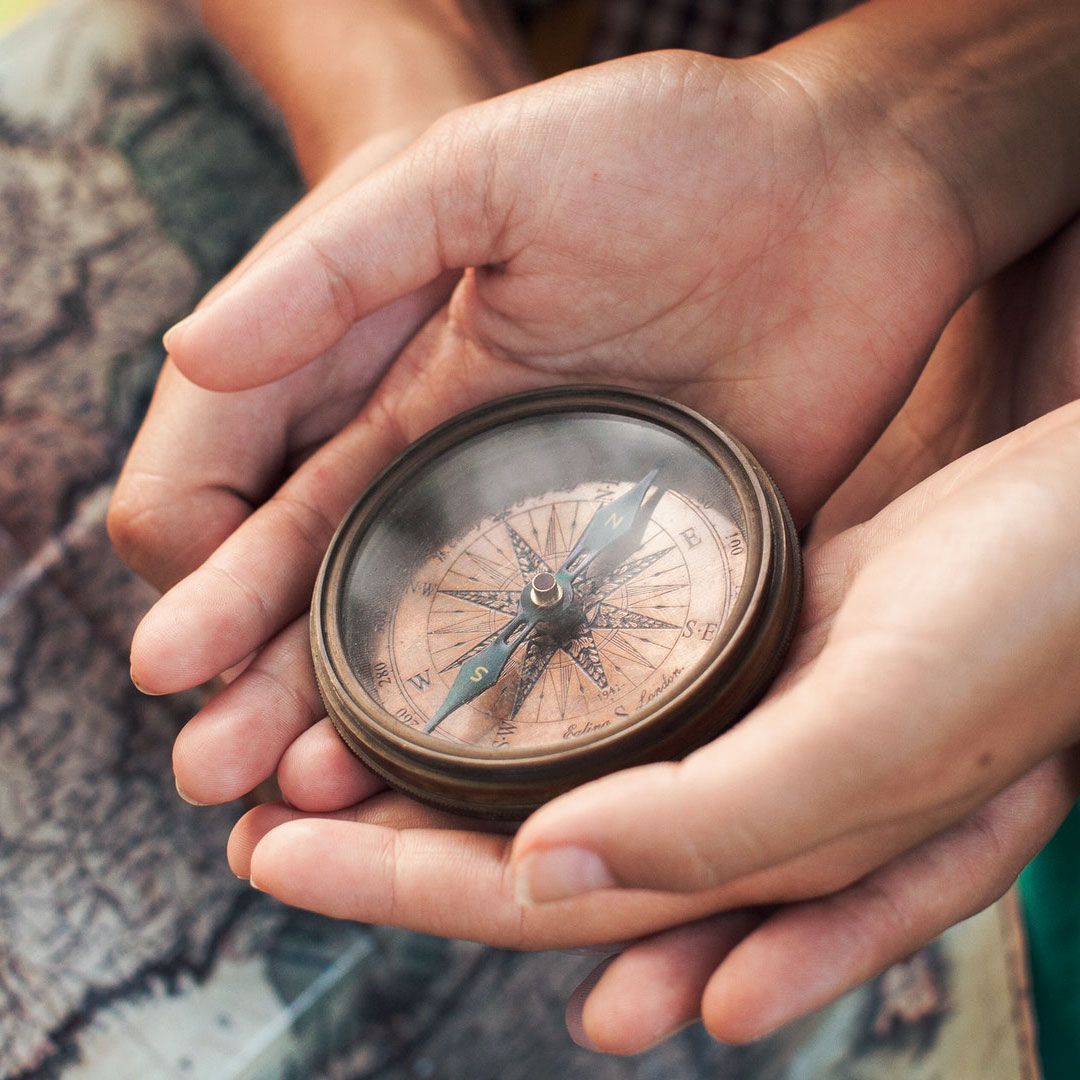 Hands cupping a vintage compass. Another pair of hands supports them from underneath.